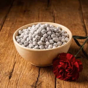 A cream-colored ceramic bowl of handcrafted Carnation incense, resting on an oak surface next to a deep red carnation bloom.